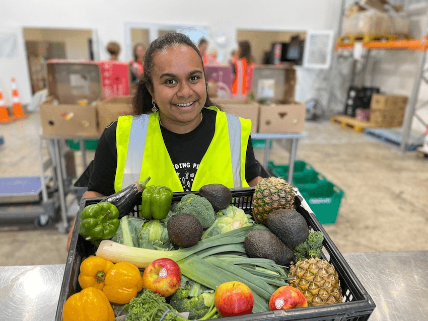 A volunteer in a fluoro vest hold s a box of donated pineapples, capsicum, leeks, apples and avocado.