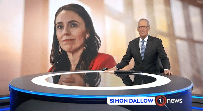 Simon Dallow sits at the 1News desk, an image of Jacinda Ardern is in the background