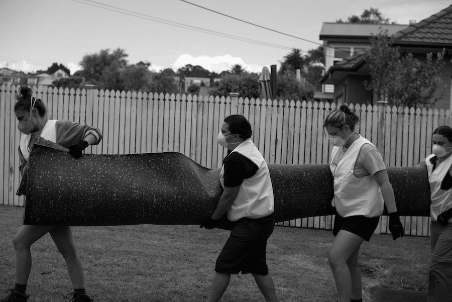 Four women in protective gear carry a rolled-up piece of wet carpet