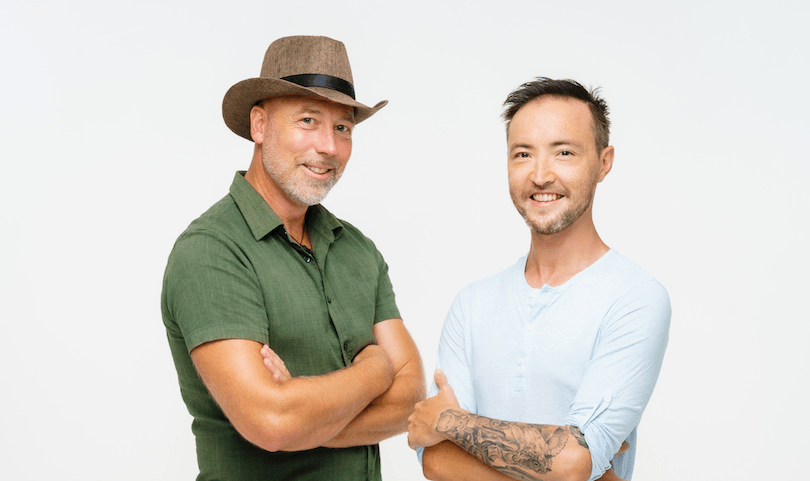 Two men, one wearing a hat, smile with their arms folded at the camera