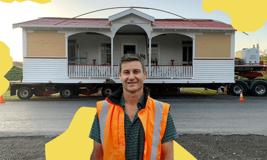 Clarke Gayford smiles while wearing an orange hi-vis vest and standing in front of a villa that's on the back of a truck