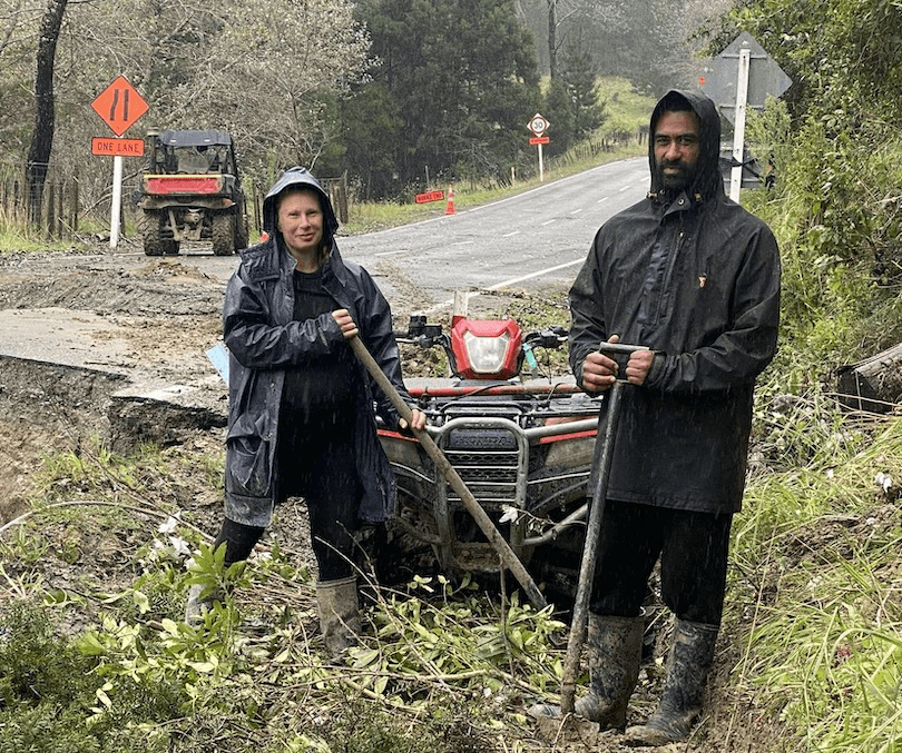 two Tokomaru residents dressed in wet weather gear holding shovels on the side of a road amid storm debris