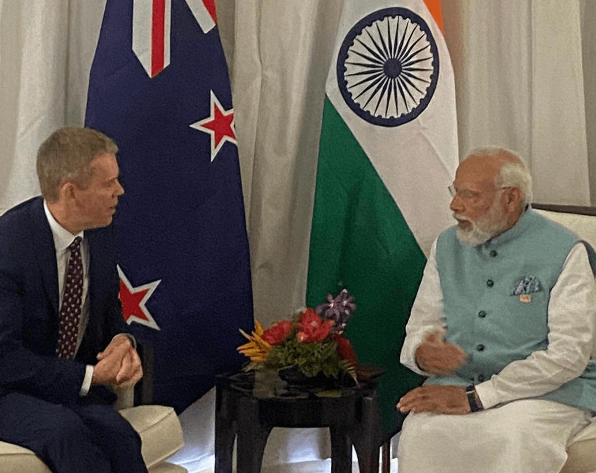with an indian and nz flag in the background, Indian priminister narendra modi with white hair and beard and a blue vest talks to chris Hipkins, a white man with light brownish reddish hair. the photo is quite dim