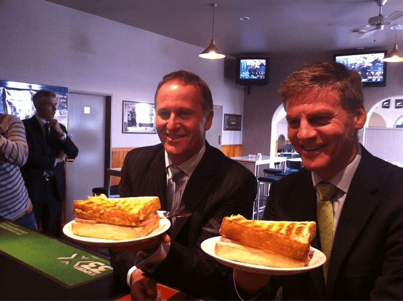John Key and Bill English grin while each holding plates with larger than normal toasted sandwiches. They look to be in a sports bar.