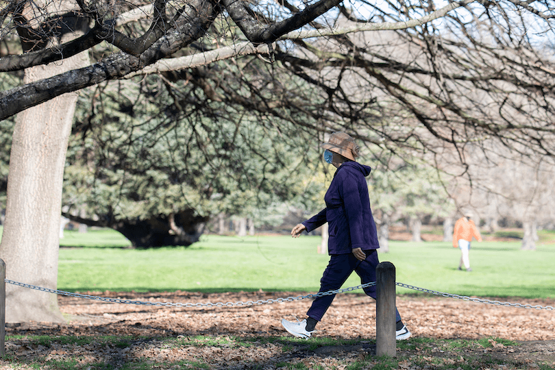 an older women wearing a hat and a face mask walking against a park backdrop