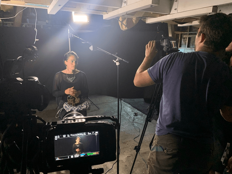a Māori woman sits on a chair, she is being filmed by a man with a camera