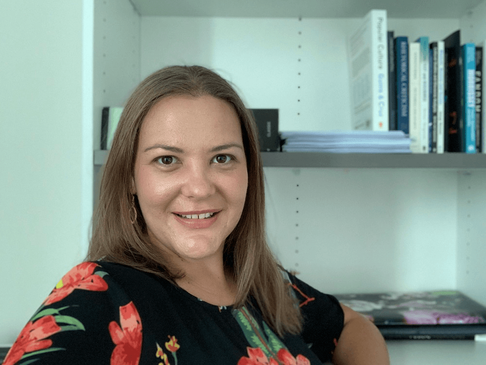 a woman with straight hair, glowy skin, and a floral shirt sitting in front of a bookshelf with a slight smile