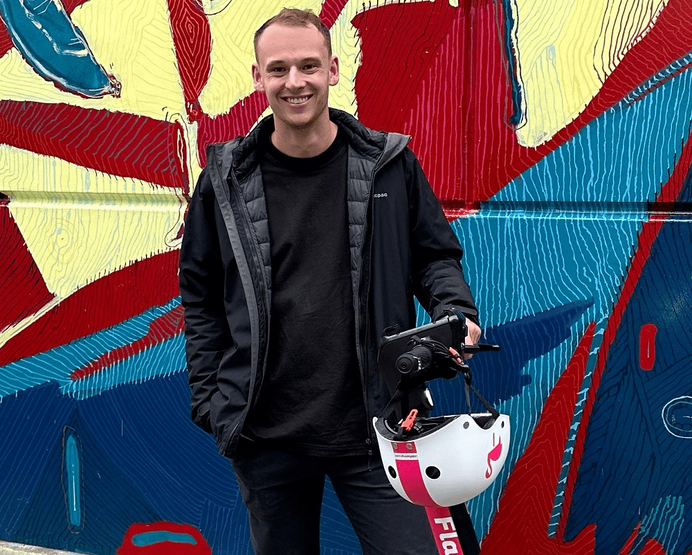 a white man in his early thirties wearing a puffa jacket stands in front of a colourful wall holding an escooter that is pink