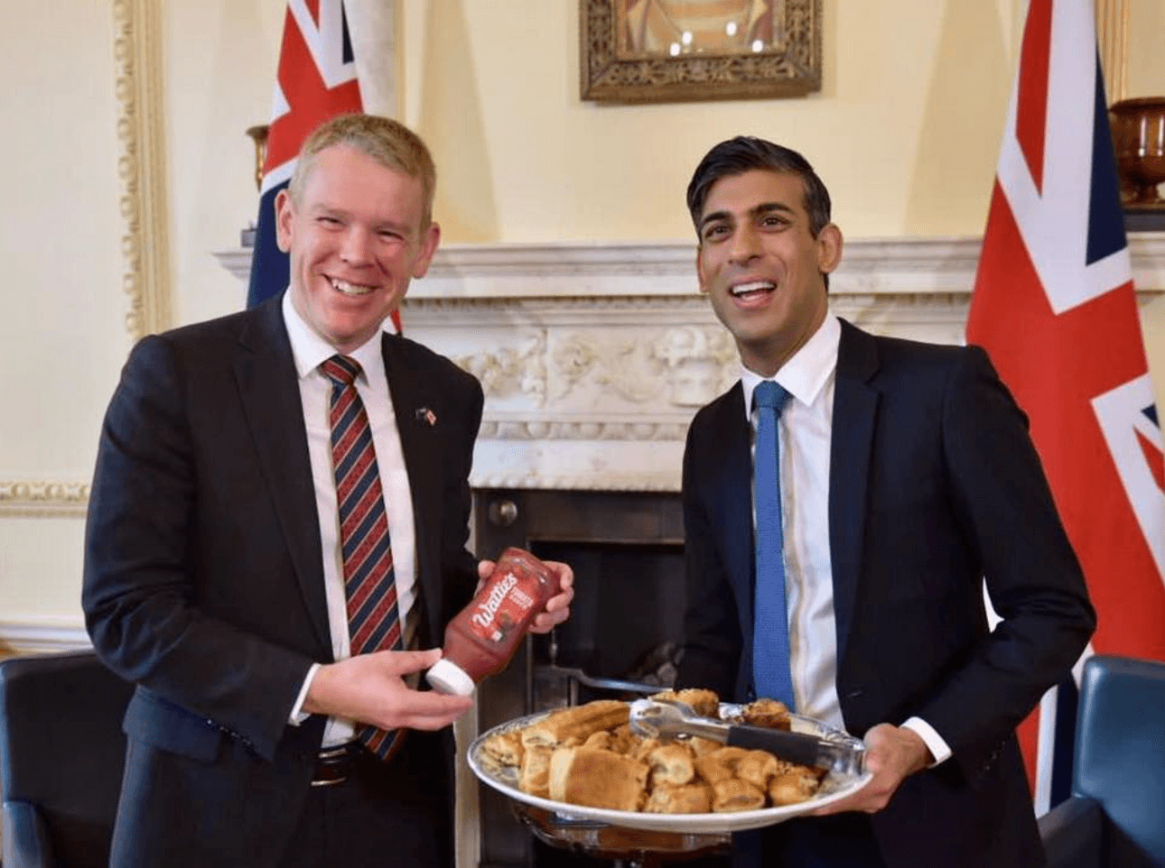 Chris Hipkins smiles with a bottle of Wattie's tomato sauce while standing next to UK prime minister Rishi Sunak. Sunak is holding a large silver platter of sausage rolls.
