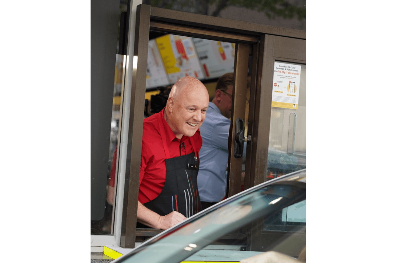 National party leader Christopher Luxon wears a red polo and a black apron as he leans out the window of a McDonald's drive through to talk to a customer..