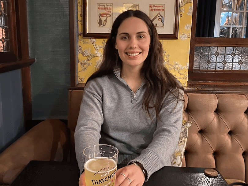 A smiling young woman with long brown hair sits in a booth seat in a typical UK pub, nursing a pint