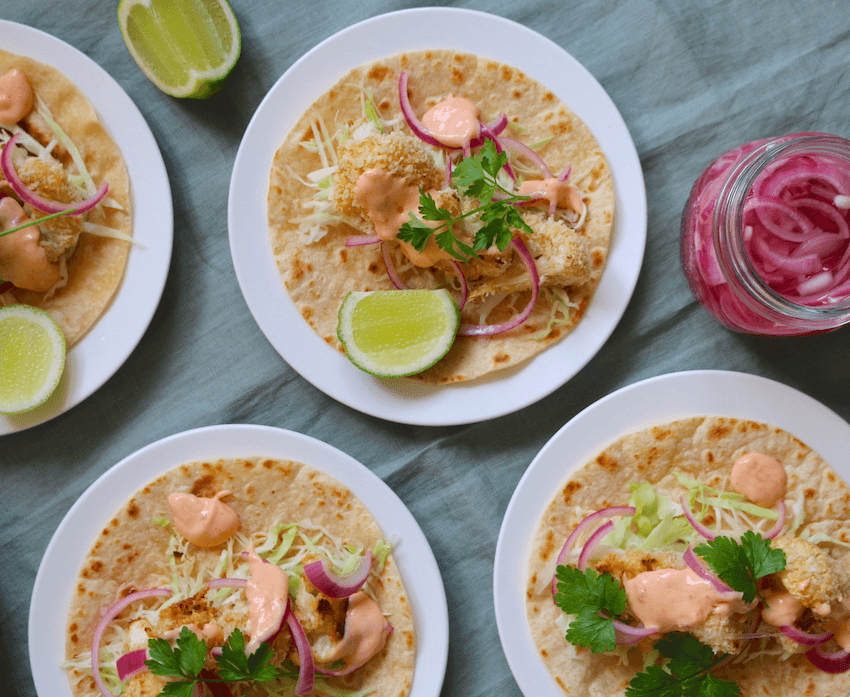 Plates of cauliflower tacos with lime wedges and pickled red onions on a grey tablecloth.