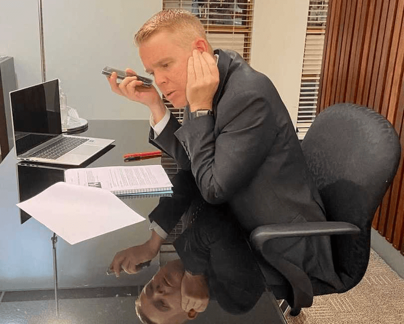 Chris Hipkins holding a phone, with his hand on his other ear. Notes and a laptop are in front of him