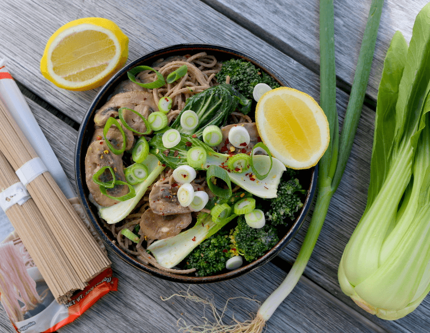 A bowl of soba noodles with broccoli, bok choy, mushrooms and spring onion on a wooden bench.