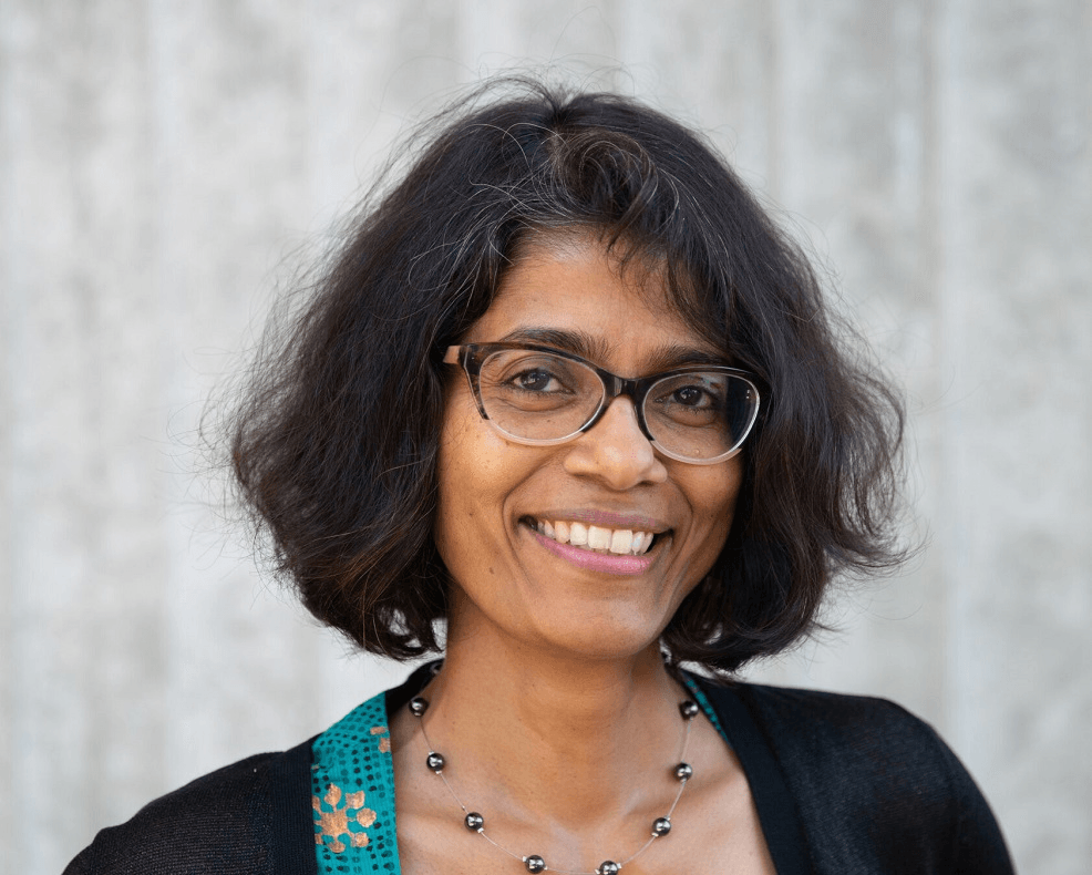 a brown skinned woman with a bobbed haircut, glasses, a big smile, and a black cardigan with a colourful top underneath