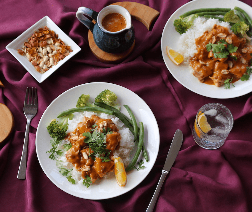 Two plates of rice topped with satay chicken. Each is served with green beans and brocolli. The plates are served on a dark purple tablecloth.