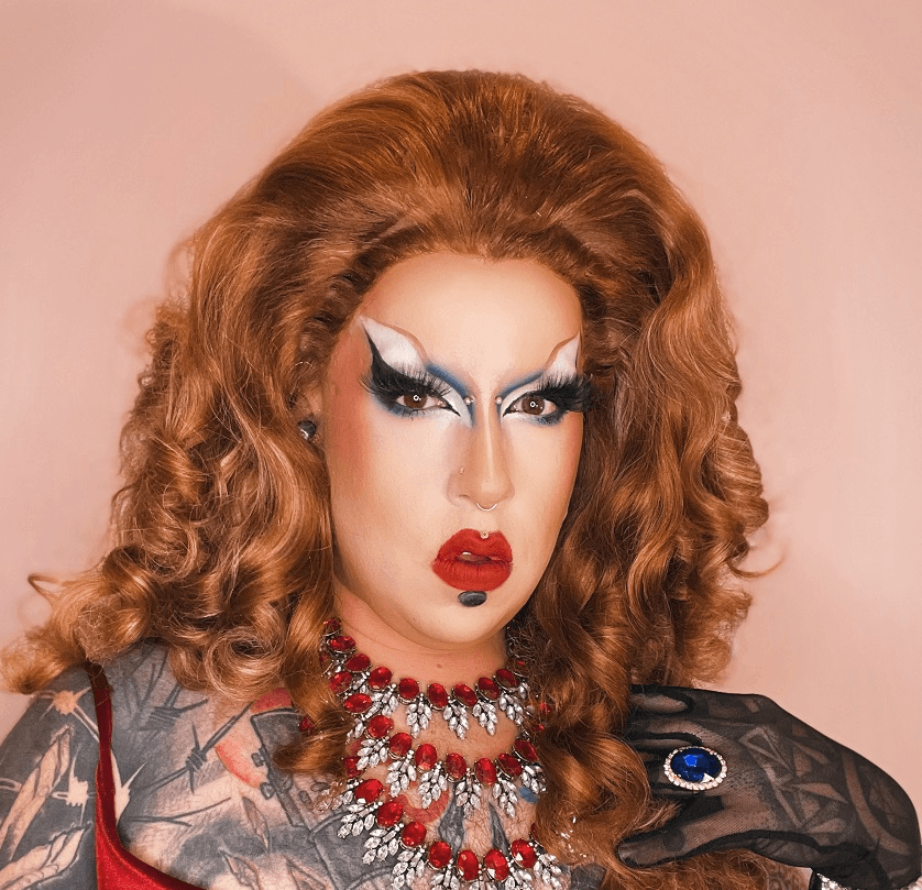 a very glamourous drag performor whith an auburn wig, red dress over lots of tattoos, sparkly red and siver and blue jewellery, bright red jeweller and cool toned dramatic white and blue eye makeup. the performer is light-skinned