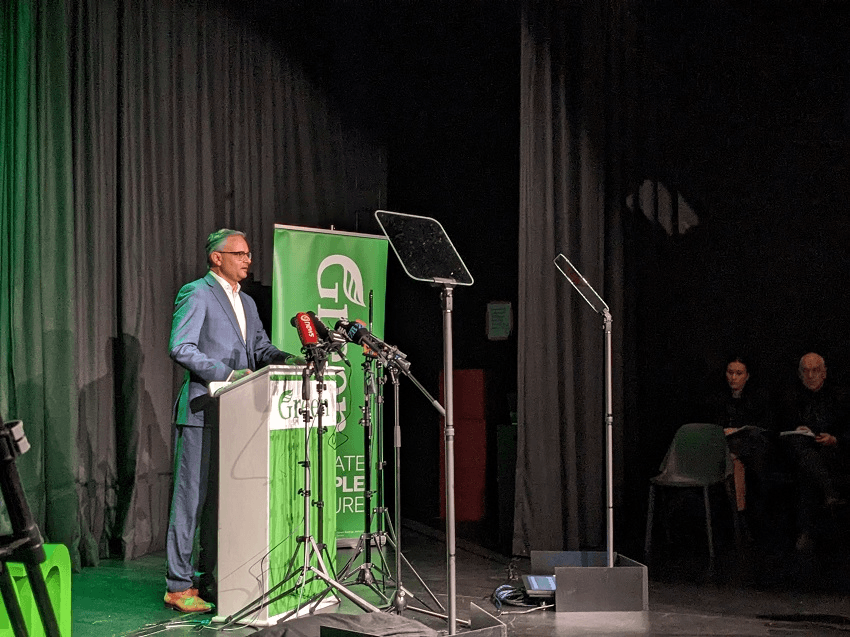 james shaw wearing a blue suit and white shirt stands at a lectern decorated with the green logo and lots of microphones around