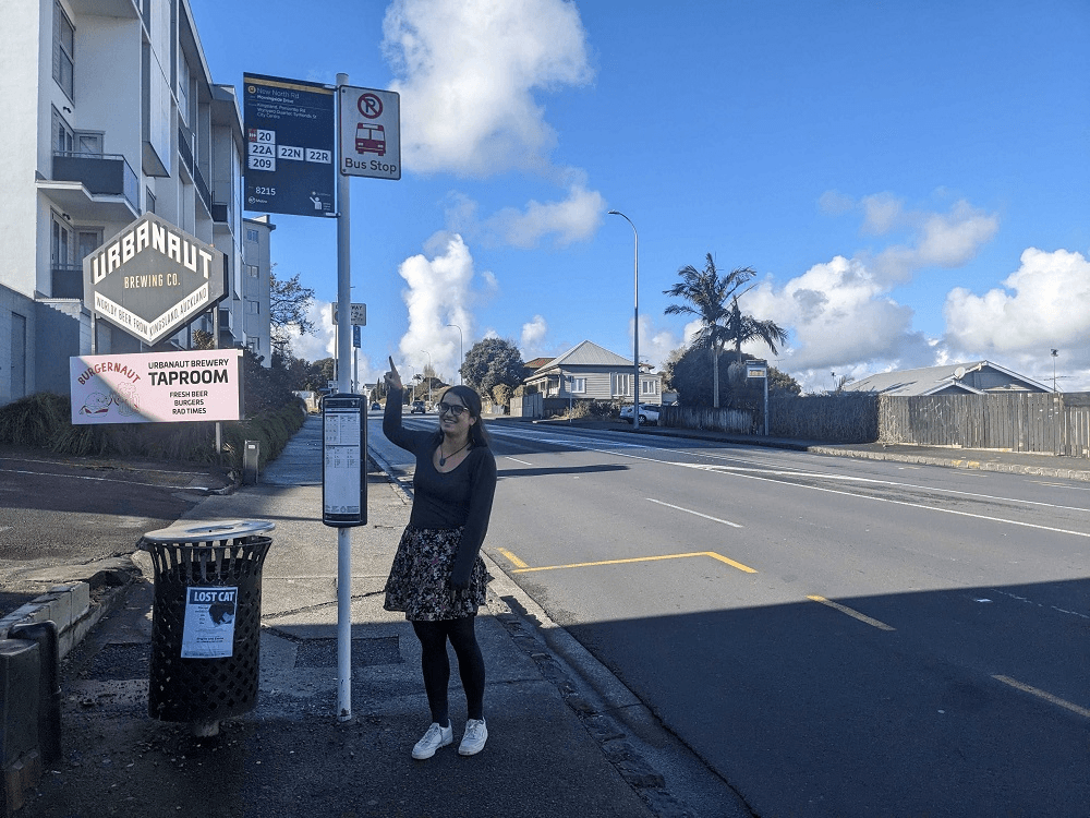 a woman with dark shortish hair and light brown skin pointing at a bus stop on a sunny day