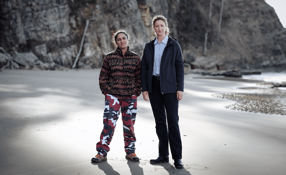 Two characters from the crime show Deadloch stand on a beach.