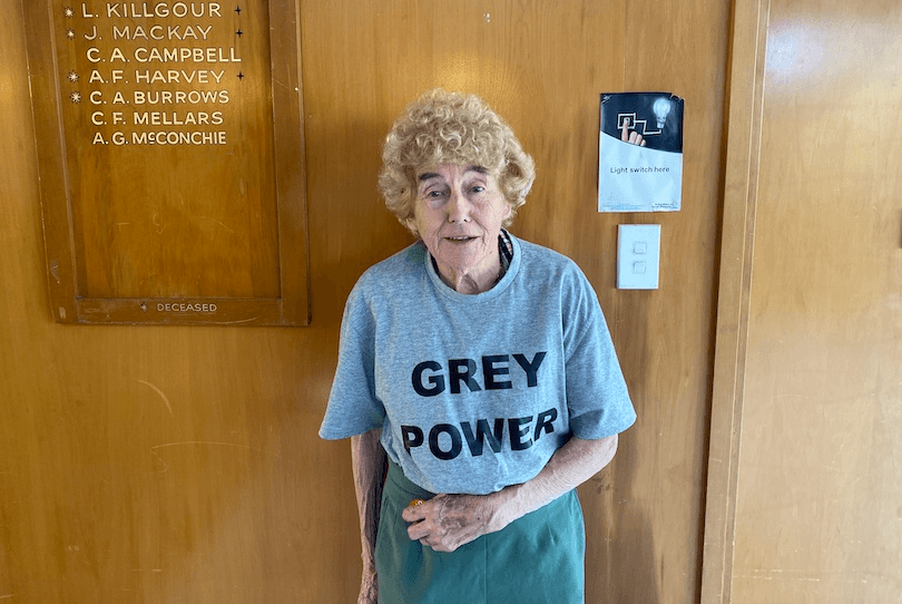 an older woman with curly grey hair, wearing a grey t-shirt emblazoned with the words GREY POWER, stands against a wooden wall, with a honours board of names behind her