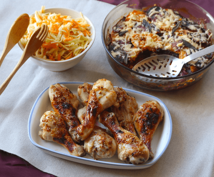 A dish of baked chicken drumsticks alongside a bowl of carrot flecked coleslaw and a large casserole dish of kūmara gratin with cheese on top. This is all presented on a beige tablecloth.