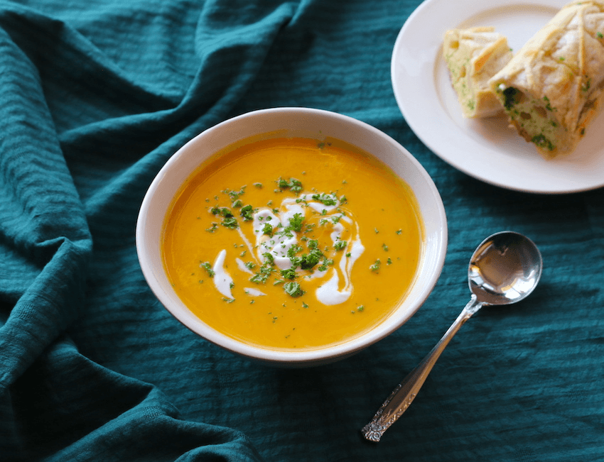 A bowl of orange kūmara soup on a dark green tablecloth with a side plate of crusty bread.
