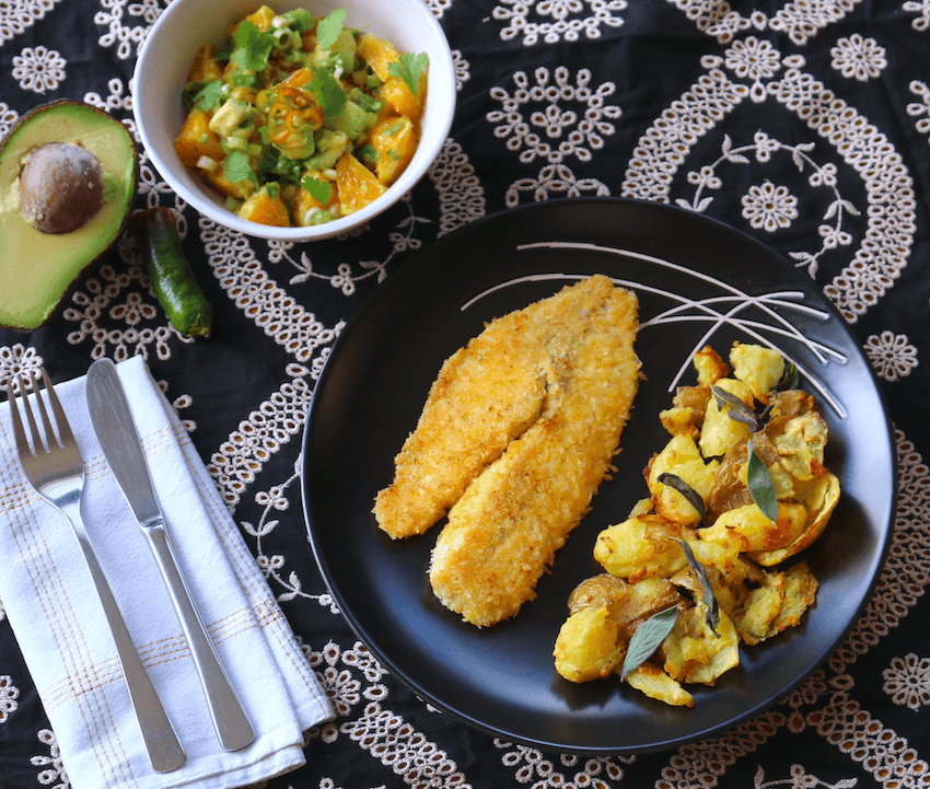 A black plate with panko crumbed fish. A white side bowl is filled with chopped mandarin segments, mint and spring onion. This si all served on a black and white patterned tablecloth.