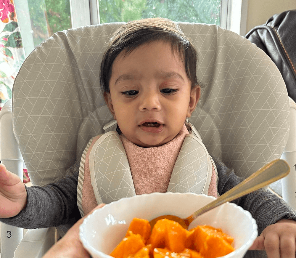 a baby in a high chair with a mango in front of her looking happy