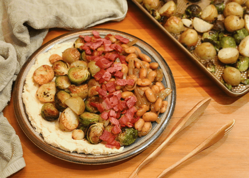 A plate topped with roasted Brussels sprouts, potatoes, and garlic cloves paired with bacon, honeyed ricotta, and creamy butter bean on a wooden bench.