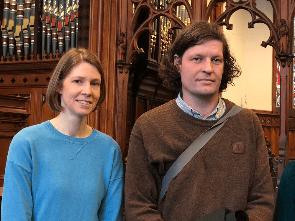a woman with short brown hair and a blue jumper standing next to a man wit rumpled brown hair and a bron jumper with a stachel over his shoulder, looking in slightly different directions, like the photographer grbbed them for the picture and they were a little bit surprised 