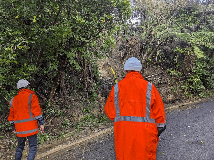 two people in orange high vis stand on a road looking at a landslide
