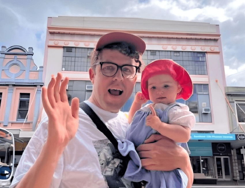 a pakeha man and baby on an auckland street looking happy and waving on a sunny day