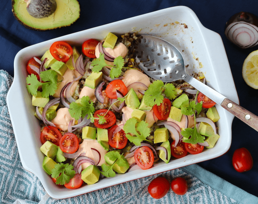 A casserole dish filled with mince, beans, and quinoa. It is topped with avocado chunks, halved cherry tomatoes, red onion slices and coriander leaves. 