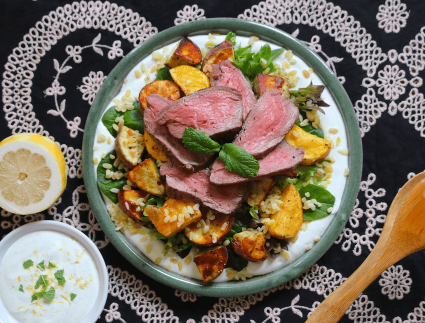 A plate piled with spinach leaves, roast vegetables, quinoa, sliced steak and a sprig of mint. Underneath the plate is an embroidered tablecloth in white and navy blue.