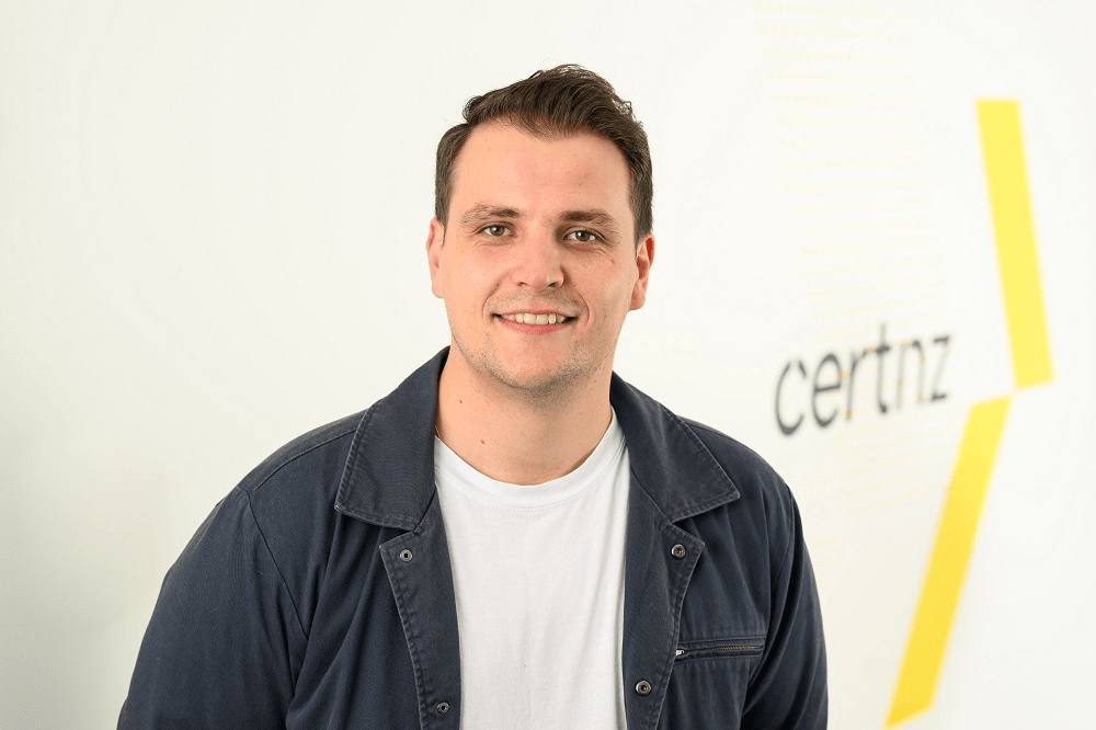 youngish man with pale skin, short brown hair, a blue jacket open to a white tshirt