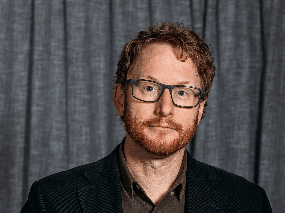 pakekha man with curly redish hair and a beard wearing black rimmed glasses and a black suit on a brey curtain background