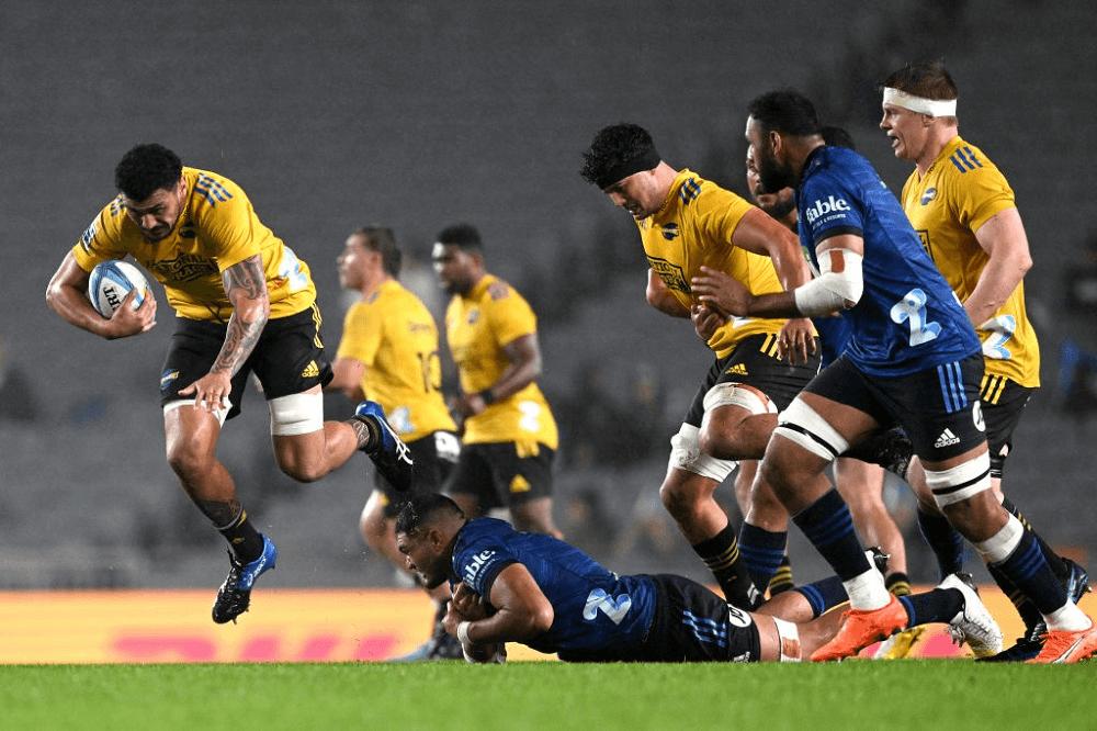 a dynamic shot of a man with a rugby ball in the air with other players in yellow and blue uniforms poised behind him