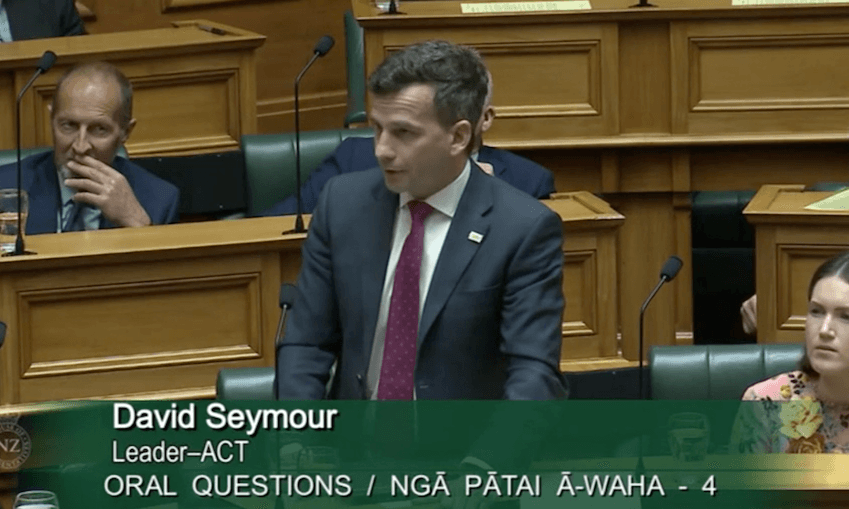 a screenshot of parliament house with david seymour standing at his desk speaking and wearing a pink tie