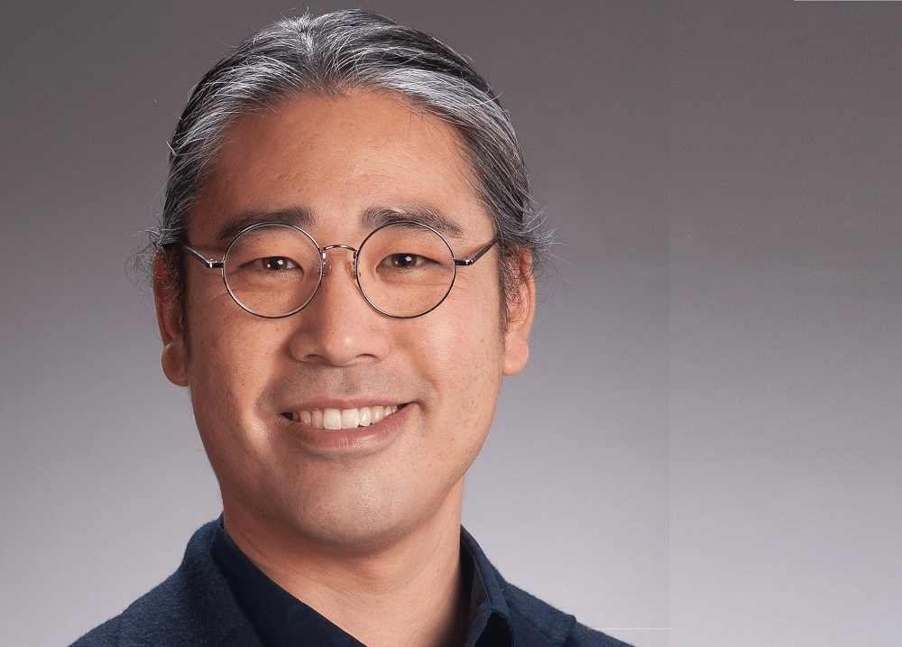 an asian man with round glasses and grey streaked hair tied in a ponytail looking at the camera with a wry smil
