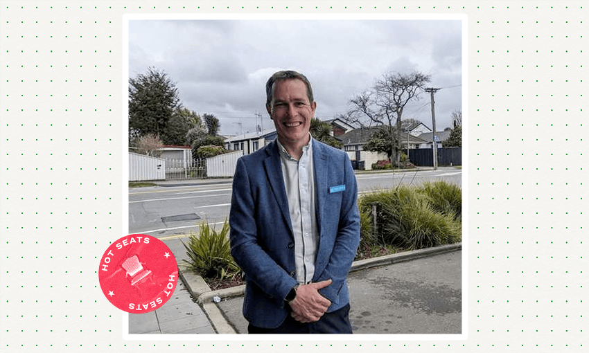 a white man in a blue suit smiling with a road and some bushes in the background