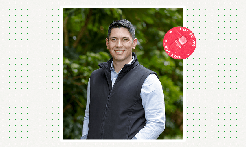 a youngish man on a green outdoors background wearing a black vest and blue shirt and smiling 