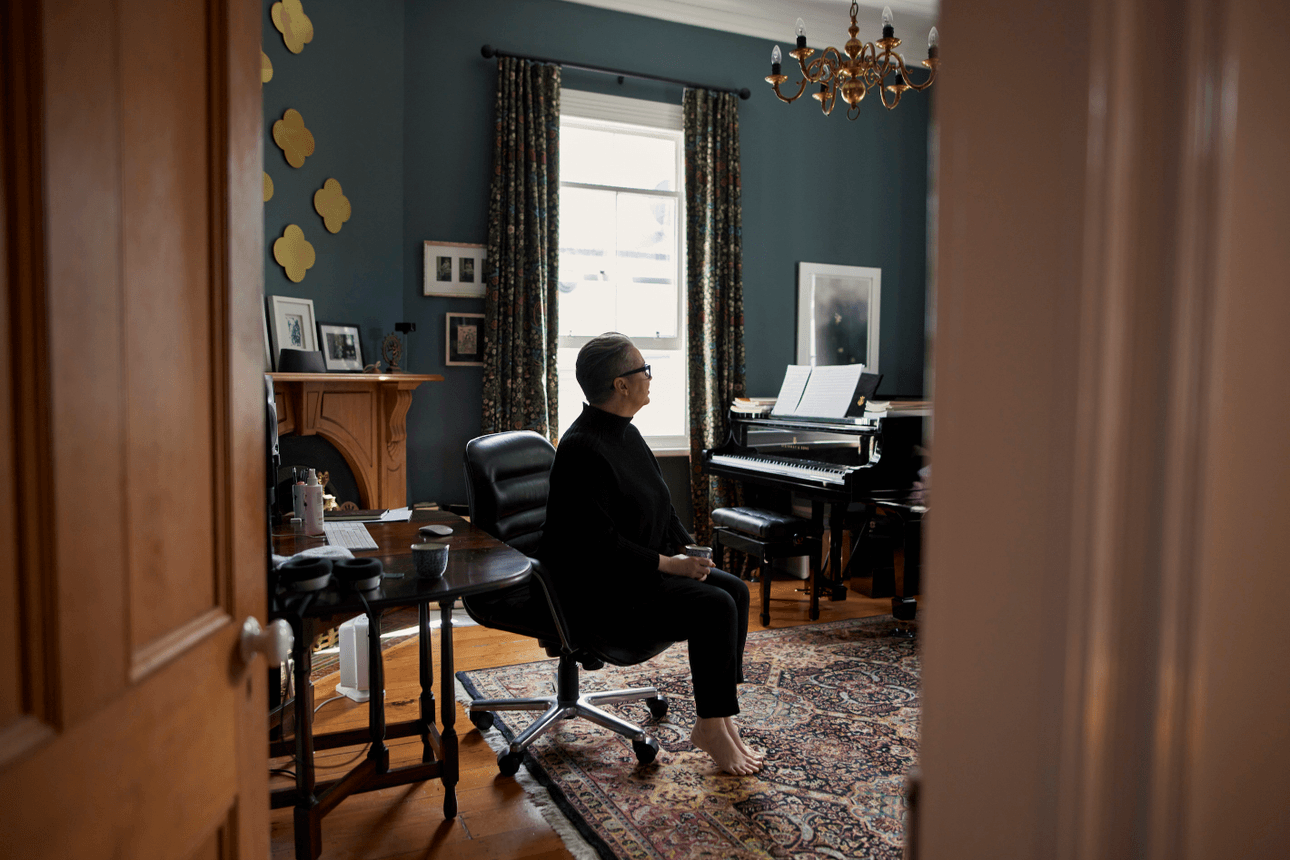 A wide shot of composer Victoria Kelly sitting in a music room in her home. The photo is shot through and framed by the doorway, and she is looking away from the camera. A Max Gimblett is visible on the wall, and a grand piano sits in the room.