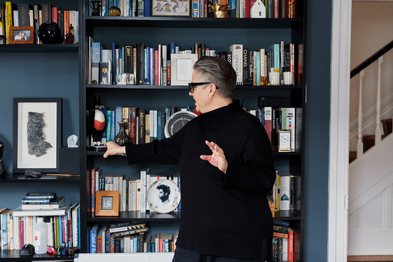 Composer Victoria Kelly stands in front of a tall bookshelf, gesturing towards a ceramic crow figurine.
