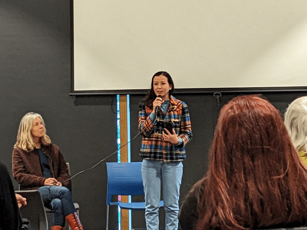 a woman in jeans and a coat speaking in front of an audience