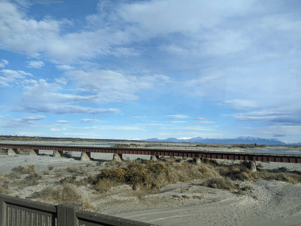 a gravelly tangled braided river bed, with a rail bridge and mountains in the background 