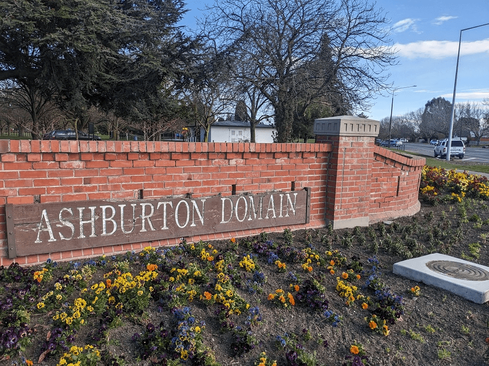 a blue sky and sunny road and a brick wall with some gardens saying "ashburton domain"