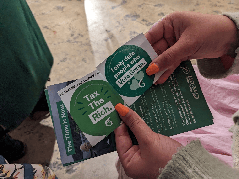 a hand with horange manicured nails holding stickers saying "tax the rich" and "I only date people who vote Green"
