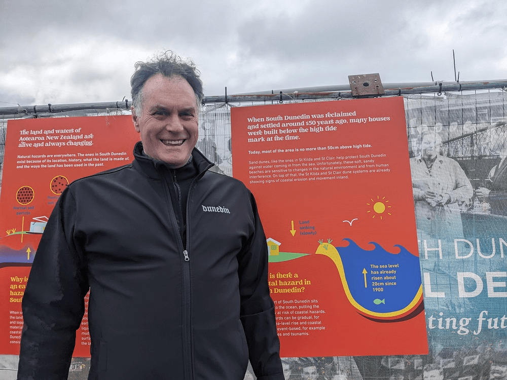 an older white man with slightly mussed dark hair and a black jacket that says Dunedin