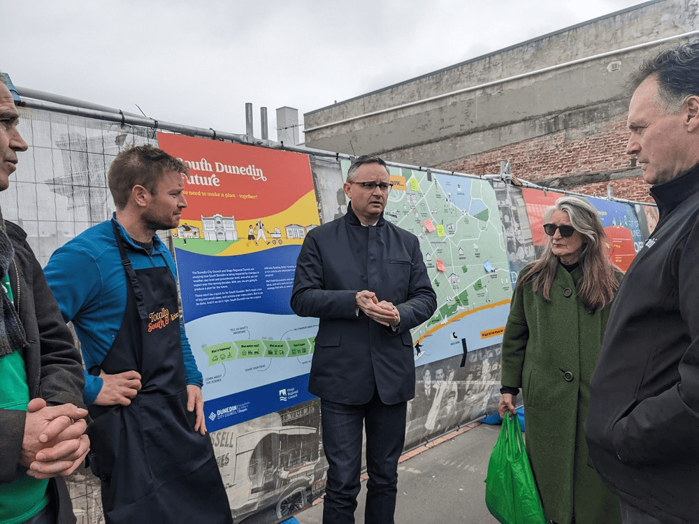 james shaw, an older man with small glasses and dark jeans with a blazer, talking to a man in a blue shirt, a woman in a green coat, and standing in front of signs about climate change in south dunedin